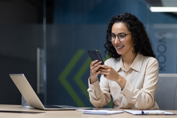 Wall Mural - Businesswoman using smartphone while working at laptop in modern office. Smiling woman checking messages, balancing technology and work. Modern office environment with technology emphasis.