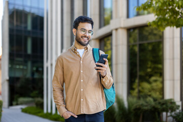 Wall Mural - Hispanic student standing outside modern university campus building, holding smartphone, smiling. Man carrying backpack, enjoying sunny day. Concept technology, education, campus life, connectivity.