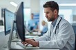 © Yuliia - young male doctor focused on his computer screen while working at his desk in a busy clinic.