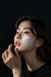 © Vadym - Korean model woman applies white cream on face in beauty studio promo. Dark background highlights facial features. Hand applying cream. Model wears black shirt, gazes directly at camera.