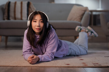 Wall Mural - Young beautiful Asian woman with headphones lying on floor in living room