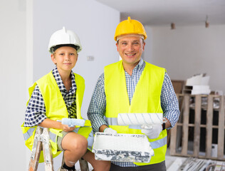 Wall Mural - Portrait of cheerful boy and his dad in yellow vests and hardhats holding paint rollers and looking at camera in construction site.