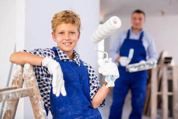 Wall Mural - Portrait of positive young boy in blue overall and hardhat holding paint roller. Repair works in apartment.