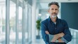 © mashimara - Portrait of a confident senior businessman with glasses and grey hair, standing with arms crossed in a modern office setting.