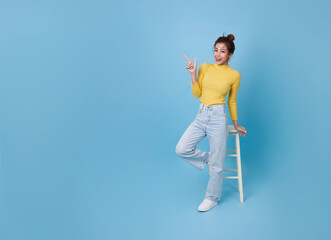 Attractive Asian teen girl sitting on chair and relax hand pointing finger isolated on blue copy space background. People lifestyle concept.