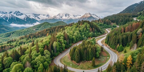 Canvas Print - A breathtaking aerial view of a winding mountain road surrounded by lush green forests and snow-capped peaks