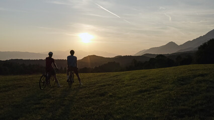 Two road cyclists silhouettes celebrating a ride finish with a high five greeting, on amazing mountain view at sunset, aerial panorama shot.