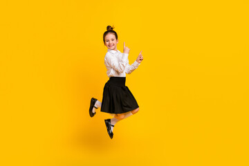 Poster - Full size photo of cute schoolgirl jump point fingers empty space wear uniform isolated on yellow color background