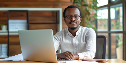 Wall Mural - A man wearing glasses sits at a desk with a laptop open in front of him. He is focused on his work, possibly typing or browsing the internet. Concept of productivity and concentration