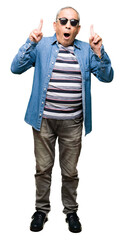 Canvas Print - Handsome senior man wearing denim jacket and sunglasses amazed and surprised looking up and pointing with fingers and raised arms.