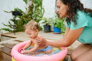 Mother bathing her baby in a small inflatable pool on a wooden deck on a sunny day