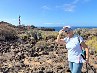 Sticker - Outdoor activity in nature for a carefree sporty senior woman walking along a rocky beach in direction to a lighthouse enjoying healthy lifestyle and freedom in good sunny day with blue sky