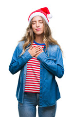 Wall Mural - Beautiful young blonde woman wearing christmas hat over isolated background smiling with hands on chest with closed eyes and grateful gesture on face. Health concept.