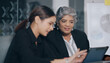 © Raushan_films - Indian older aged boss women use tablet touch pad look screen talk new deal plan share ideas with teen girl indoor job office young adult expert female old elder lady staff do work day task work place