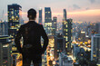 © sergign - A man in a suit stands with his back to the camera, looking out over a bustling city skyline at sunset, symbolizing ambition and future aspirations.