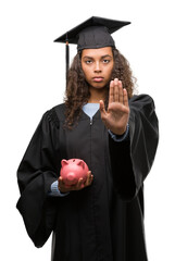 Canvas Print - Young hispanic woman wearing graduation uniform holding piggy bank with open hand doing stop sign with serious and confident expression, defense gesture