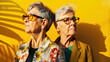 © chekart - A candid portrait of two elderly lesbians against a yellow wall. Two gray-haired women with short hair and glasses are standing next to each other and looking in different directions