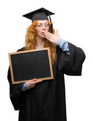 Wall Mural - Young redhead student woman wearing graduated uniform holding blackboard cover mouth with hand shocked with shame for mistake, expression of fear, scared in silence, secret concept