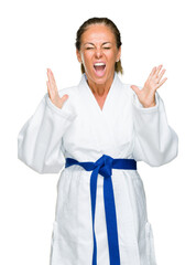 Wall Mural - Middle age adult woman wearing karate kimono uniform over isolated background celebrating mad and crazy for success with arms raised and closed eyes screaming excited. Winner concept