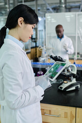 Aufkleber - Young Chinese female scientist in gloves and lab coat pointing at screen of tablet while studying new virus or strain in laboratory