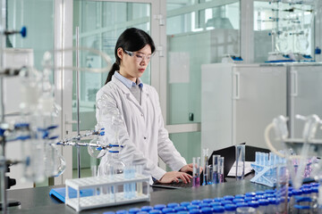 Aufkleber - Young Asian female scientist in lab coat and protective eyeglasses looking at laptop screen and typing on keyboard during network