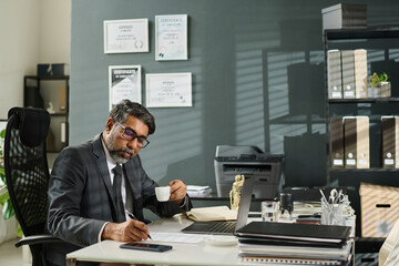 Wall Mural - Mature male lawyer with cup of coffee in hand bending over workplace with laptop and other office supplies and signing contract