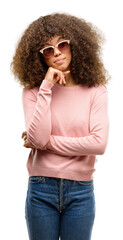 Poster - African american woman wearing pink sunglasses with hand on chin thinking about question, pensive expression. Smiling with thoughtful face. Doubt concept.