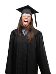 Wall Mural - Young hispanic woman wearing graduated cap and uniform sticking tongue out happy with funny expression. Emotion concept.