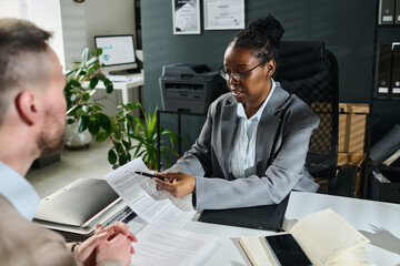 Wall Mural - Confident African American businesswoman or lawyer pointing at title of paper document while explaining male client its main points