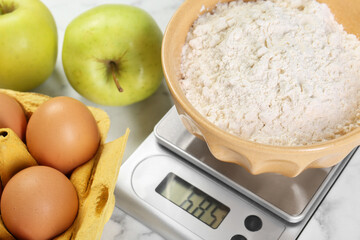 Wall Mural - Kitchen scale with bowl of flour, eggs and apples on white table, closeup