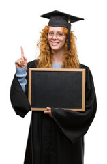 Poster - Young redhead student woman wearing graduated uniform holding blackboard surprised with an idea or question pointing finger with happy face, number one