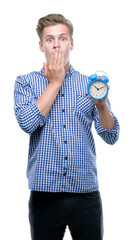 Poster - Young handsome blond man holding alarm clock cover mouth with hand shocked with shame for mistake, expression of fear, scared in silence, secret concept