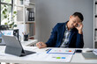 © Wasana - A man in a blue jacket is sitting at a desk with a laptop and a stack of papers