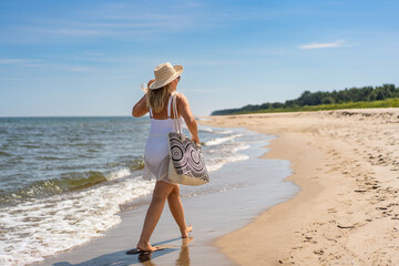 Wall Mural - Summer vacation by Baltic Sea. Beautiful young woman in white swimsuit and white pareo and beige sun hat with beach bag walking by sea on sandy beach on beautiful sunny day. Back view