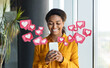 © Prostock-studio - Black woman smiles as she looks at her phone. Heart icons surround her, representing notifications or likes on a social media app. The background features a simple office setting.