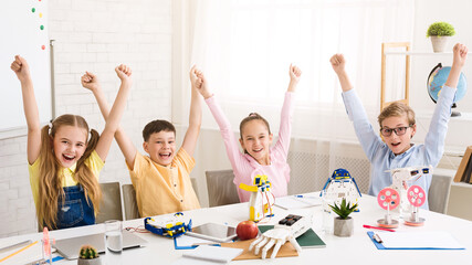 Aufkleber - Four children sit at a table in a classroom, arms raised in the air, celebrating a successful robotics project. They are surrounded by their work, including robots, tools, and other materials