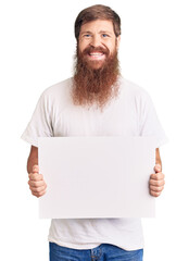 Wall Mural - Handsome young red head man with long beard holding blank empty banner looking positive and happy standing and smiling with a confident smile showing teeth