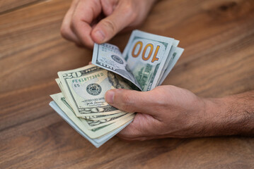 Poster - Male company accountant holds dollar banknotes of different denominations carefully counting cash for weekly income report at end of week sitting at wooden table in office
