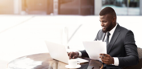 Entrepreneurship. Cheerful Business Guy Using Laptop Working With Papers Sitting In Outdoor Cafe In City. Copy Space