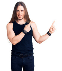 Young adult man with long hair wearing goth style with black clothes smiling and looking at the camera pointing with two hands and fingers to the side.