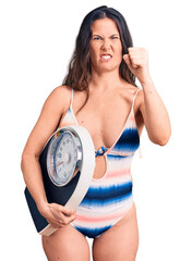 Wall Mural - Young beautiful brunette woman wearing swimwear holding weighing machine annoyed and frustrated shouting with anger, yelling crazy with anger and hand raised