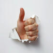 © Denniro - Hand giving thumbs up through torn paper. Close-up of a hand giving a thumbs up gesture, breaking through a torn piece of white paper, symbolizing approval and positivity