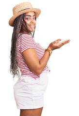 Poster - Young african american woman with braids wearing summer hat pointing aside with hands open palms showing copy space, presenting advertisement smiling excited happy