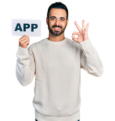 Wall Mural - Young hispanic man with beard holding app banner doing ok sign with fingers, smiling friendly gesturing excellent symbol