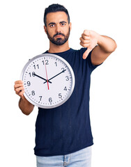 Poster - Young hispanic man holding big clock with angry face, negative sign showing dislike with thumbs down, rejection concept