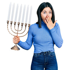 Canvas Print - Young brunette woman with blue eyes holding menorah hanukkah jewish candle covering mouth with hand, shocked and afraid for mistake. surprised expression