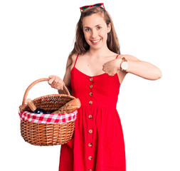 Canvas Print - Young beautiful blonde woman holding picnic basket pointing finger to one self smiling happy and proud