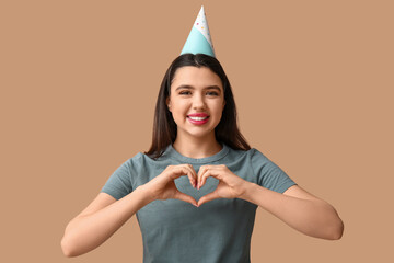 Poster - Beautiful young woman in party hat showing heart gesture on brown background