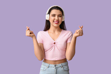 Poster - Beautiful young woman in modern headphones showing heart gesture on lilac background