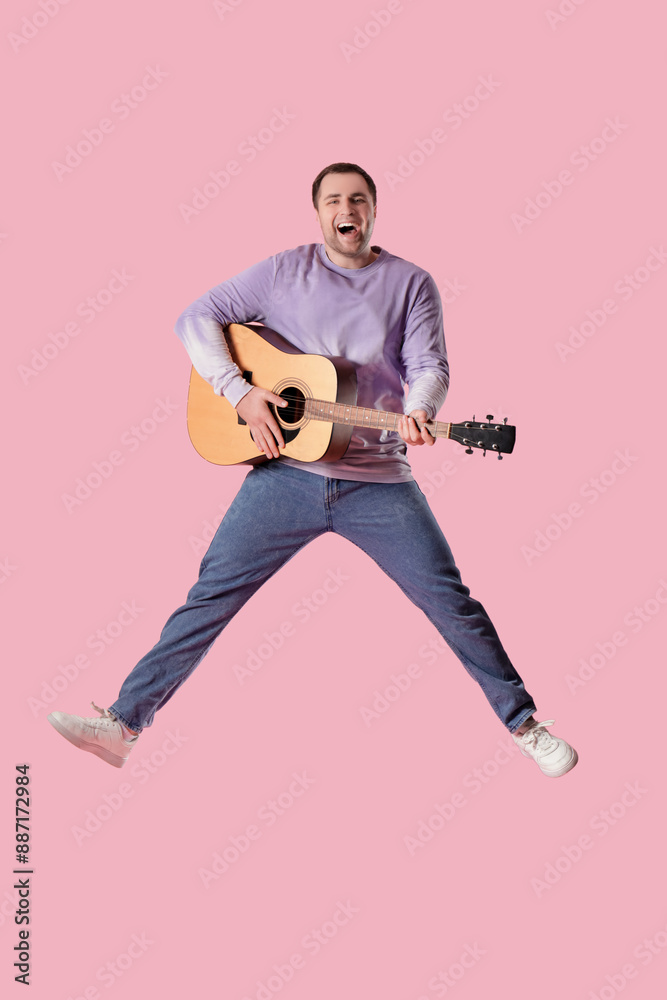 Young happy man with guitar jumping on pink background